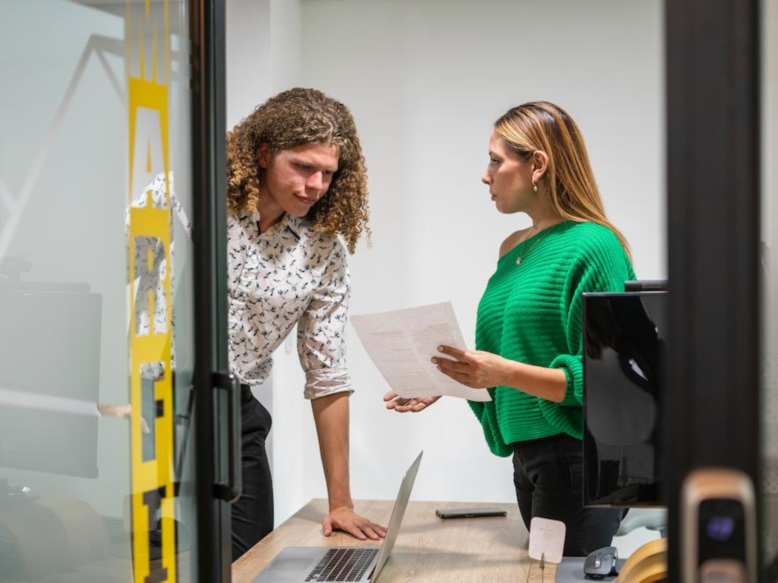 To kollegaer i samtale bak lukket dør, der én holder et dokument – illustrerer betydningen av konfidensialitet og beskyttelse av forretningshemmeligheter i arbeidsmiljøet.