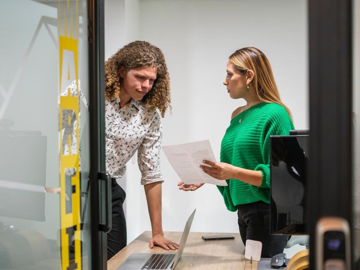 To kollegaer i samtale bak lukket dør, der én holder et dokument – illustrerer betydningen av konfidensialitet og beskyttelse av forretningshemmeligheter i arbeidsmiljøet.